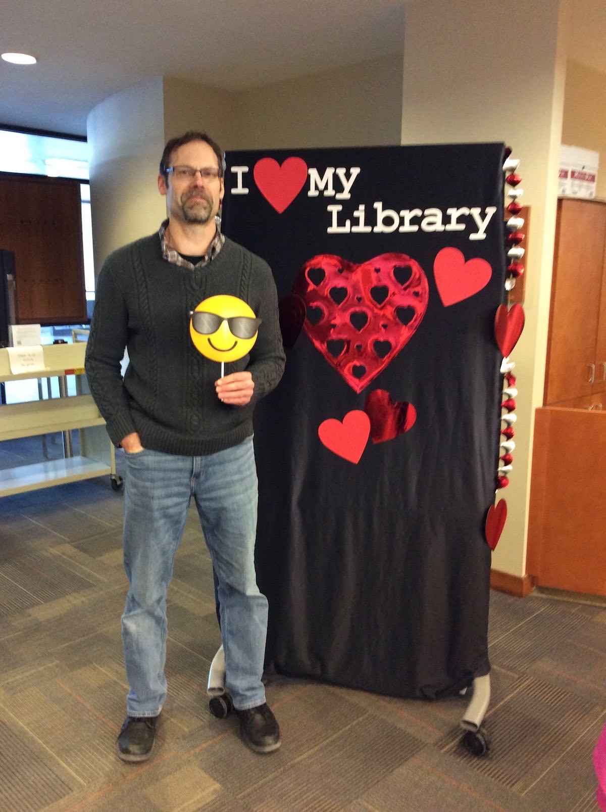 Librarian Michael Shires stands in front of the I love my Library backdrop holding a prop of a smiling sunglasses emoji up in front of his torso
